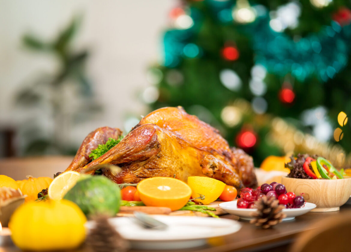 A holiday-themed potluck table featuring a roasted turkey with colorful fruits and side dishes, set in front of a decorated Christmas tree.